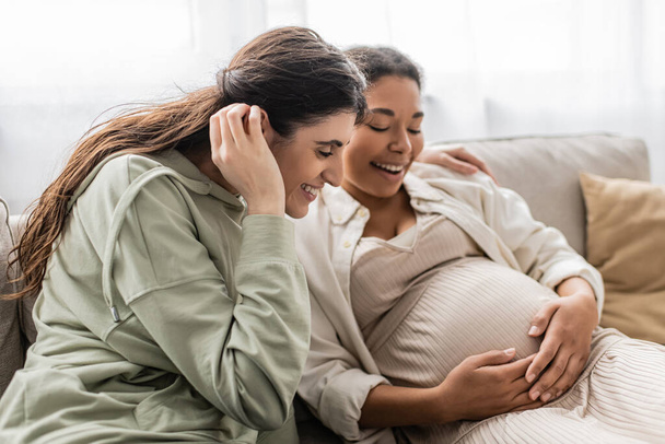 happy lesbian woman smiling while hugging pregnant multiracial wife while sitting on sofa  - Zdjęcie, obraz