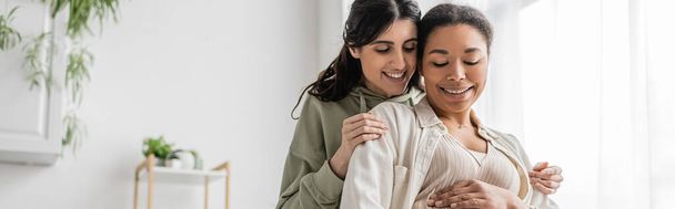 happy lesbian woman smiling while hugging cheerful wife in living room, banner  - Photo, Image