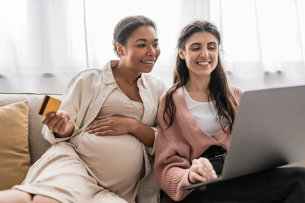 pregnant multiracial woman holding credit card while doing online shopping with happy lesbian partner  - Foto, afbeelding