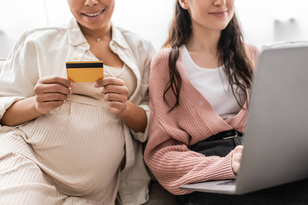 przycięty widok ciężarnej wielorasowej kobiety posiadającej kartę kredytową podczas robienia zakupów online z wesołym partnerem lesbijki  - Zdjęcie, obraz