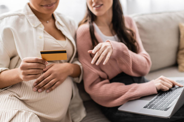 partial view of pregnant multiracial woman holding credit card while doing online shopping with lesbian partner  - Fotografie, Obrázek