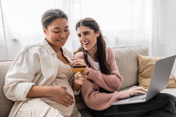feliz embarazada y multirracial mujer sosteniendo tarjeta de crédito mientras hace compras en línea con alegre pareja lesbiana  - Foto, Imagen