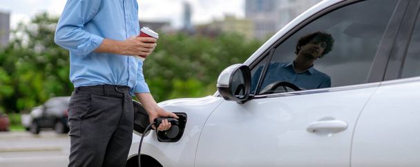Progressive eco-friendly concept of parking EV car at public electric-powered charging station in city with blur background of businessman leaning on recharging-electric vehicle with coffee. - Zdjęcie, obraz