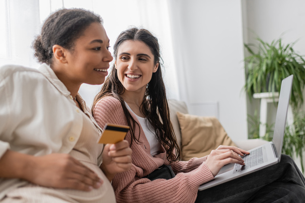 happy lesbian woman looking at pregnant multiracial partner while doing online shopping together  - Photo, Image