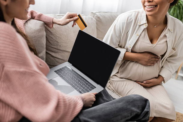 cropped view of cheerful lesbian woman holding credit card near pregnant multiracial partner while doing online shopping together  - Фото, изображение