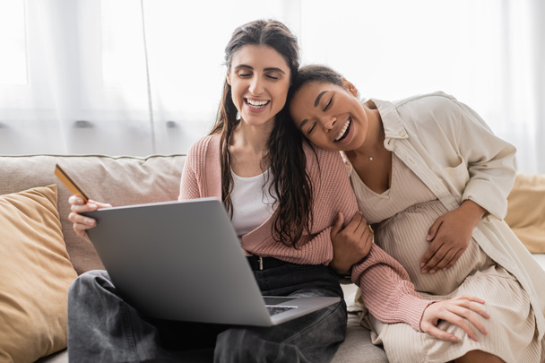 happy lesbian woman holding credit card while doing online shopping near pregnant multiracial partner  - 写真・画像