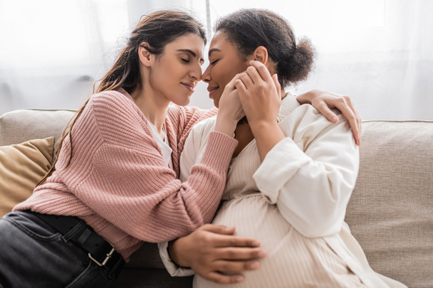 happy lesbian woman holding hands with pregnant multiracial partner and sitting on couch  - Fotó, kép