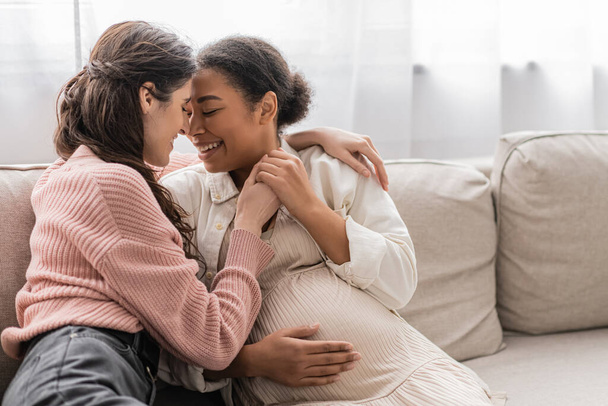 positive lesbian woman hugging pregnant multiracial partner and sitting on couch  - Фото, изображение