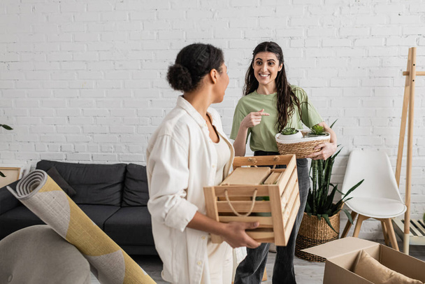 happy lesbian woman pointing at wicker basket with plants near multiracial girlfriend during relocation to new house  - Φωτογραφία, εικόνα