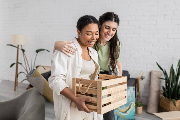 overjoyed lesbian woman hugging multiracial girlfriend holding wooden box during relocation to new house  - Fotoğraf, Görsel