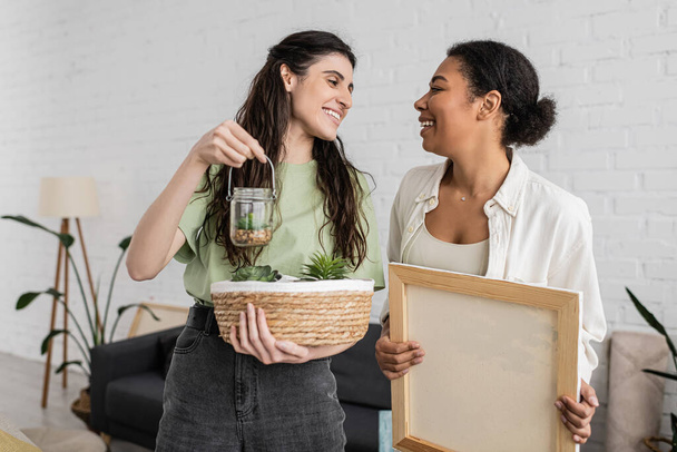 cheerful lesbian woman holding wicker basket and glass jar with plants next to happy multiracial girlfriend in new house  - Photo, Image