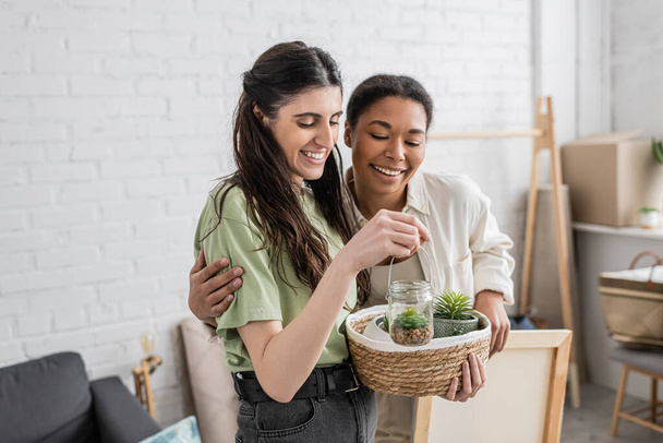 cheerful lesbian woman holding wicker basket and glass jar with plants next to positive multiracial girlfriend in new house  - Photo, Image
