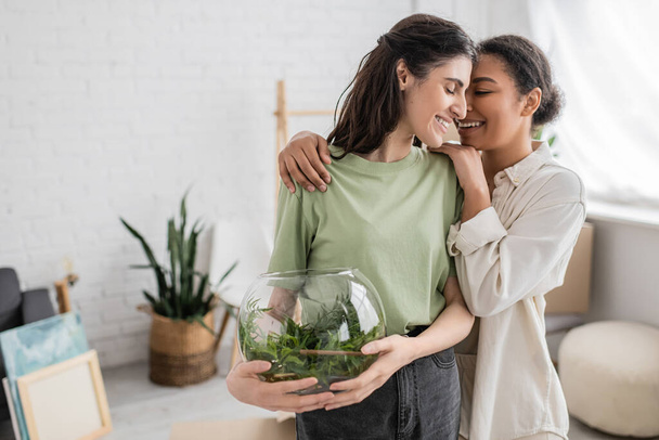 positive multiracial woman hugging lesbian partner with green plant in glass vase  - Foto, Bild