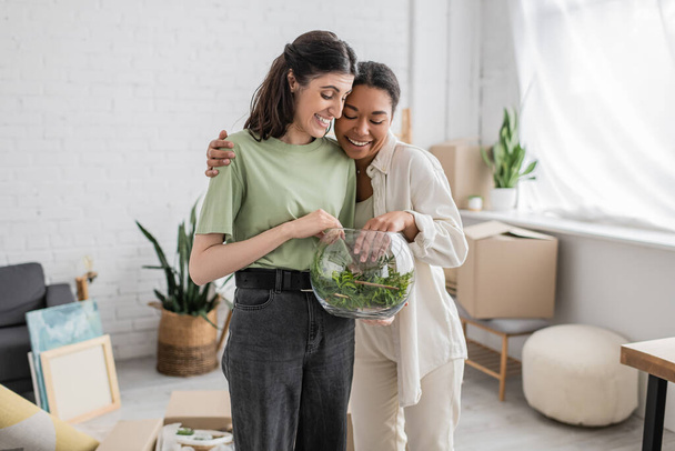overjoyed interracial and lesbian women hugging and looking at green plant in glass vase  - Foto, Imagem