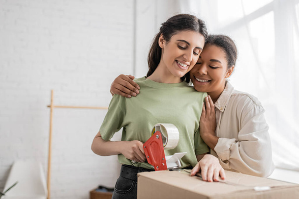 wesoły wielorasowy kobieta przytulanie szczęśliwy Lesbijki partner taping karton pudełko podczas relokacji do nowego domu  - Zdjęcie, obraz