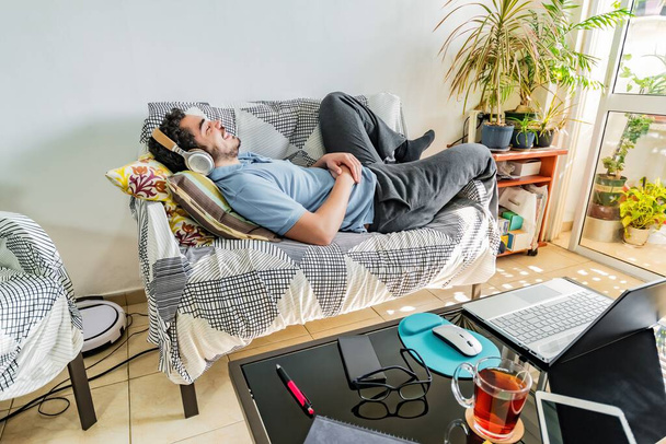 Молодой мужчина в трениках лежит дома на диване и слушает музыку в наушниках - Фото, изображение