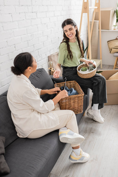 overhead view of happy interracial lesbian couple holding wicker baskets while sitting on sofa in living room  - Φωτογραφία, εικόνα