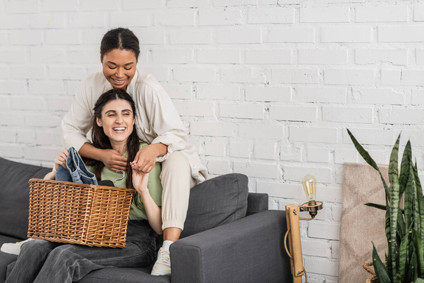 cheerful lesbian woman folding clean laundry while sitting on sofa next to multiracial girlfriend   - Photo, Image