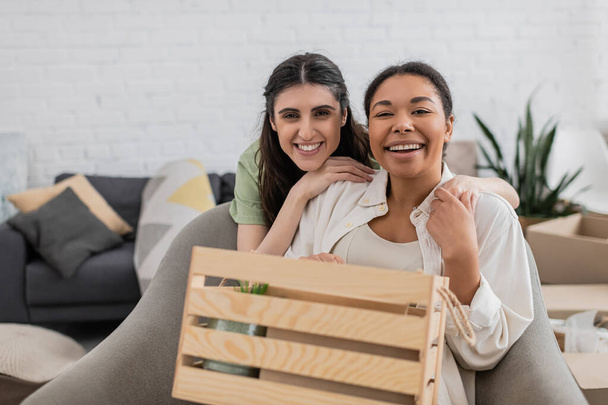 joyful lesbian woman hugging happy multiracial girlfriend near wooden box in living room - 写真・画像