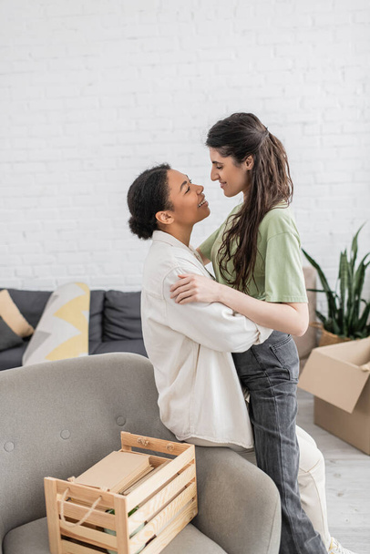 positive lesbian woman hugging happy multiracial girlfriend near wooden box in living room  - Foto, afbeelding