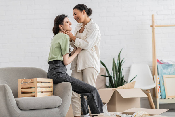 cheerful lesbian woman hugging joyful multiracial girlfriend with closed eyes in living room of new house  - Foto, Bild