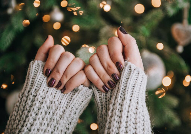 Close-up shot of female hands with painted nails in front of christmas tree with bokeh lights. - Foto, imagen