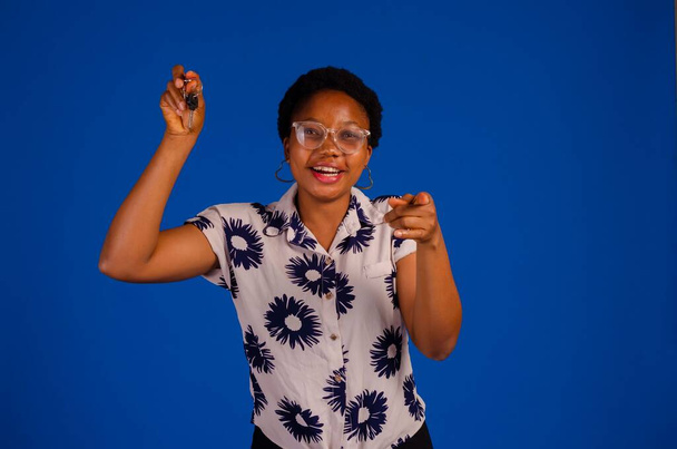 An African woman holding keys and pointing at the camera isolated on a blue background. - Valokuva, kuva