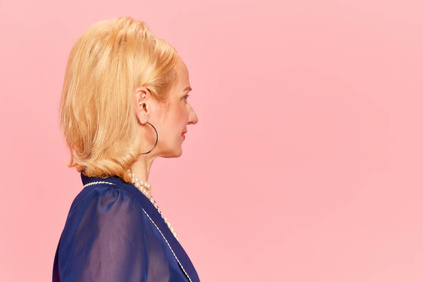 Portrait avec une jolie femme blonde avec une coupe de cheveux rétro debout et regardant sur fond de studio rose. Vue du profil, gros plan. Concept d'émotions humaines, mode, beauté, vintage, publicité - Photo, image
