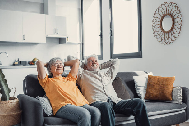 Peaceful middle aged man and woman with closed eyes relaxing on comfortable couch at home, mature family daydreaming together, grey haired wife and husband resting with hands behind head, breathing - Фото, изображение