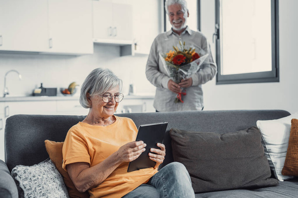Old man giving flowers at his wife sitting on the sofa at home for the San Valentines day. Pensioners enjoying surprise together. In love people having fun.  - Φωτογραφία, εικόνα