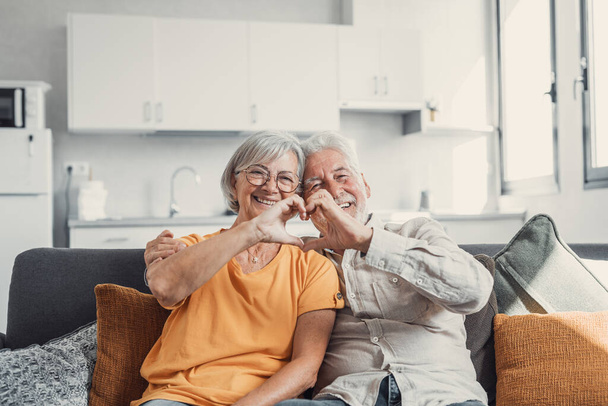 Close up portrait happy sincere middle aged elderly retired family couple making heart gesture with fingers, showing love or demonstrating sincere feelings together indoors, looking at camera. - Fotó, kép