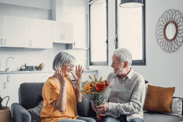 Old man giving flowers at his wife sitting on the sofa at home for the San Valentines day. Pensioners enjoying surprise together. In love people having fun.  - Foto, Bild
