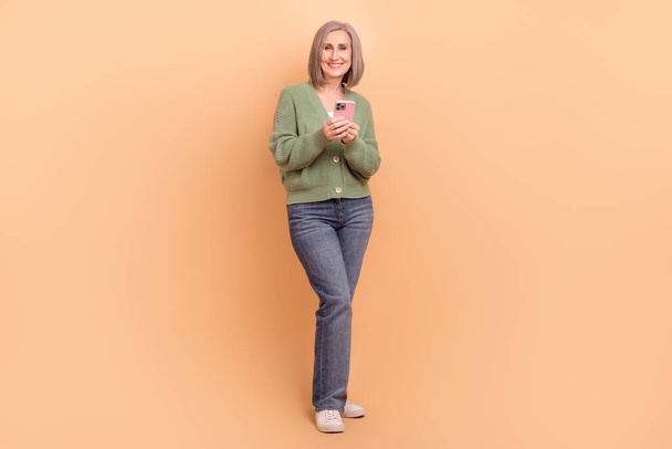 Full length cadre photo of optimistic pensioner businesswoman blogger hold phone chatting with family isolated on beige color background. - Фото, изображение