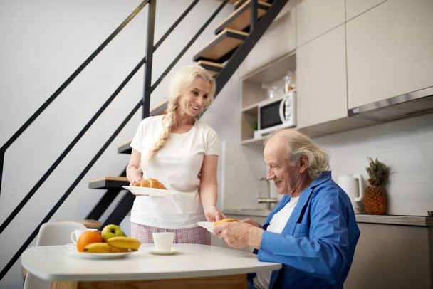 Happy senior couple, man and woman having breakfast together in kitchen, woman serving croissants with coffee. Cheerful talks. Love, relationship, family, emotions, lifestyle, support and care concept - Photo, image