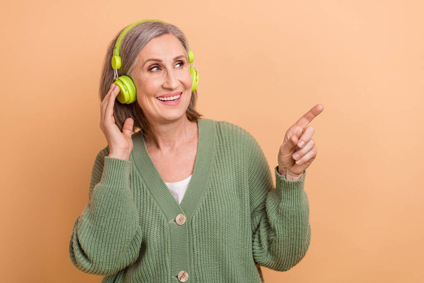 Photo of cheerful aged person arm touch headphones look direct finger empty space isolated on beige color background. - Valokuva, kuva