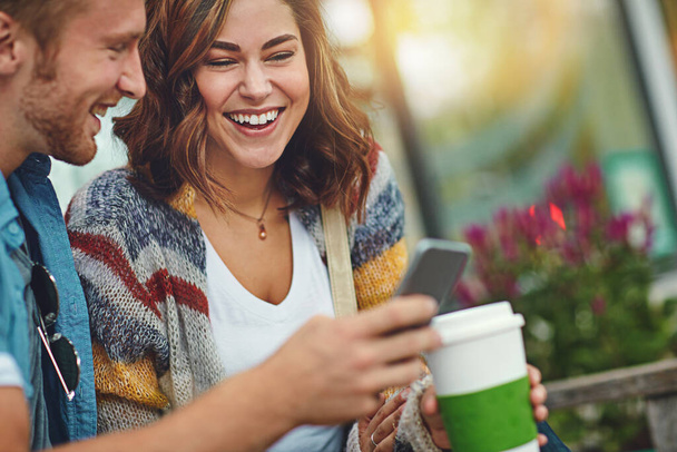 Nunca les falta el entretenimiento. una feliz pareja joven usando un teléfono inteligente mientras pasan el día en el centro de la ciudad - Foto, Imagen