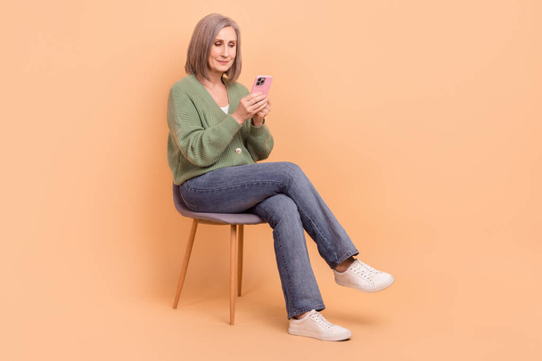 Full size photo of good mood retired person wear knit pullover look at smartphone sitting on chair isolated on pastel color background. - Fotoğraf, Görsel
