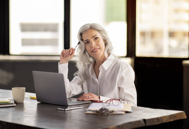 Wirtschaftskarriere. Frau mittleren Alters posiert am Laptop im modernen Büro und lächelt in die Kamera. Professionelle Personalmanagerin arbeitet am Computer am Arbeitsplatz. Headhunting und Beschäftigung - Foto, Bild