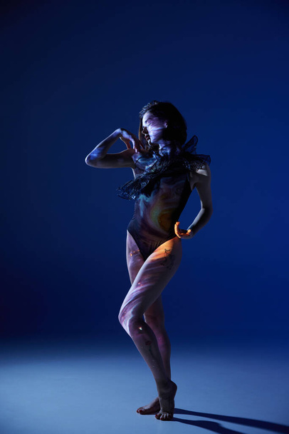 Krása ženského těla. Jedna mladá smyslná žena v čistě černém obleku, dotýkající se jejího těla na modrém pozadí studia v neonovém světle. Pojem emoce, digitální umění, móda, futurismus, umění - Fotografie, Obrázek