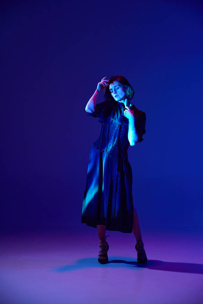 One charming woman wearing black maxi dress and heels posing at camera with peaceful facial expression over dark blue studio background in neon light. Concept of beauty, human emotions, fashion, art - Фото, изображение
