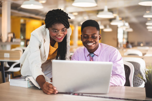 Χαμογελώντας νεαρός αφροαμερικανός επιχειρηματίας και επιχειρηματίας χρησιμοποιώντας φορητό υπολογιστή, ενώ συζητούν για το νέο σχέδιο του έργου. - Φωτογραφία, εικόνα