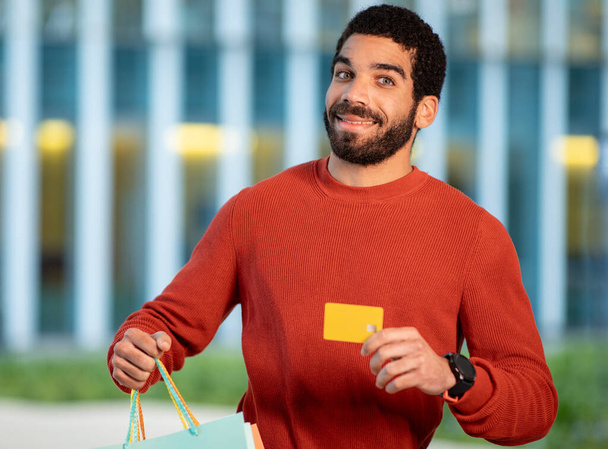 Shopping Offer. Cheerful Bearded Brunette Man Showing His Credit Card And Paper Shopper Bags, Advertising Bank Payment Service Smiling To Camera Standing Outdoors. Selective Focus - Photo, Image