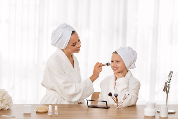Loving mother and her little daughter in bathrobes doing makeup at home, caring mom teaching her child beauty tricks, using blusher brush, happy family enjoying spending time together, copy space - Photo, Image