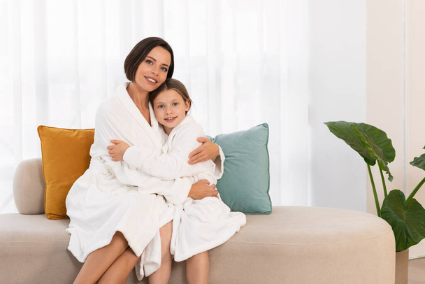 Porträt der schönen glücklichen jungen Mutter und kleinen Tochter, die in weißen Bademänteln posiert, liebevolle Mutter und ein frühes weibliches Kind, das zu Hause auf der Couch sitzt, sich umarmt und in die Kamera lächelt, Kopierraum - Foto, Bild
