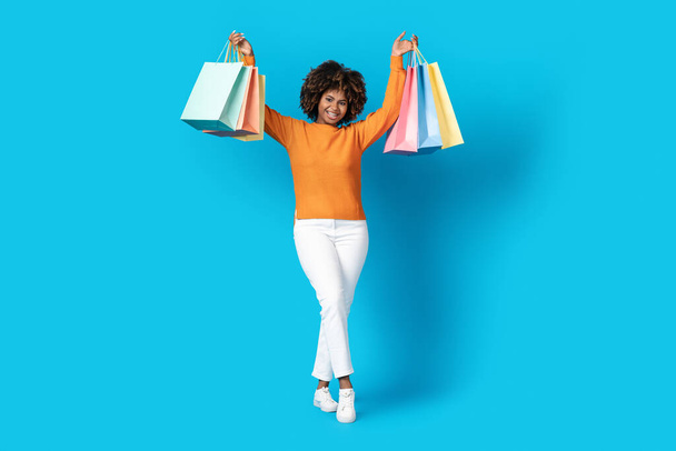 Feliz mujer africana con estilo joven con pelo tupido shopaholic mostrando bolsas de compras de colores en sus manos sobre fondo de estudio azul, disfrutando de acuerdo viernes negro, espacio de copia - Foto, imagen
