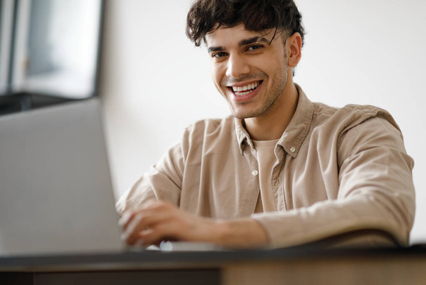 ラップトップコンピュータを使用して幸せなアラビア人現代のオフィスでテーブルに座ってオンラインで入力して作業しています。職場の屋内でカメラを構える若いビジネスマンの笑顔。選択的フォーカス - 写真・画像