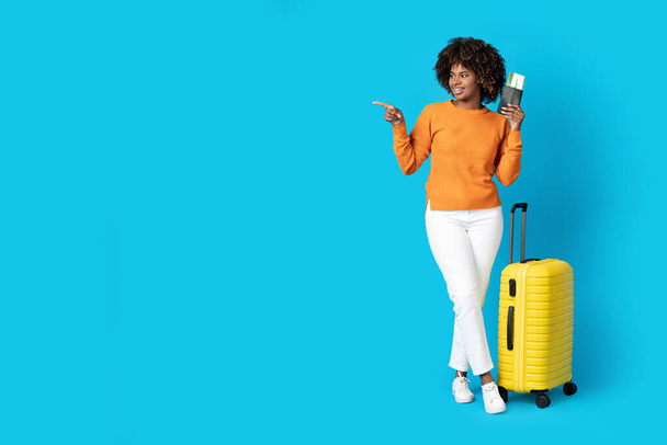 Θετική χαμογελαστή νεαρή Αφροαμερικανή που φοράει άνετο casual ντύσιμο με κίτρινες αποσκευές τουρίστριες που δείχνουν το χώρο των αντιγράφων, κρατώντας διαβατήριο και αεροπορικά εισιτήρια, δείχνοντας ωραία συμφωνία - Φωτογραφία, εικόνα