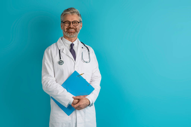 Healthcare concept. Cheerful friendly positive handsome middle aged grey-haired european doctor in workwear with stethoscope on neck and medical chart posing on blue studio background, copy space - Photo, Image