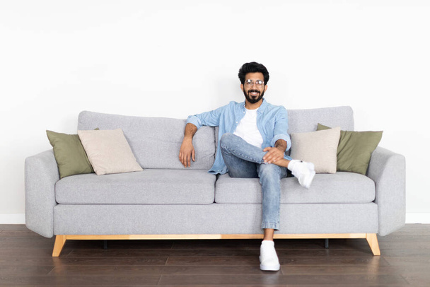 Relaxed cheerful young eastern guy wearing eyeglasses and casual comfy outfit chilling alone on couch at home, smiling at camera, copy space, enjoying his new house, resting at weekend - Foto, Bild