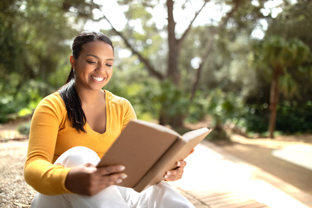 Lachende Braziliaanse studente die buiten een boek leest, zich voorbereidt op lezingen of examens terwijl ze op een bank zit in een moderne universiteitscampus of -park, kopieerruimte - Foto, afbeelding